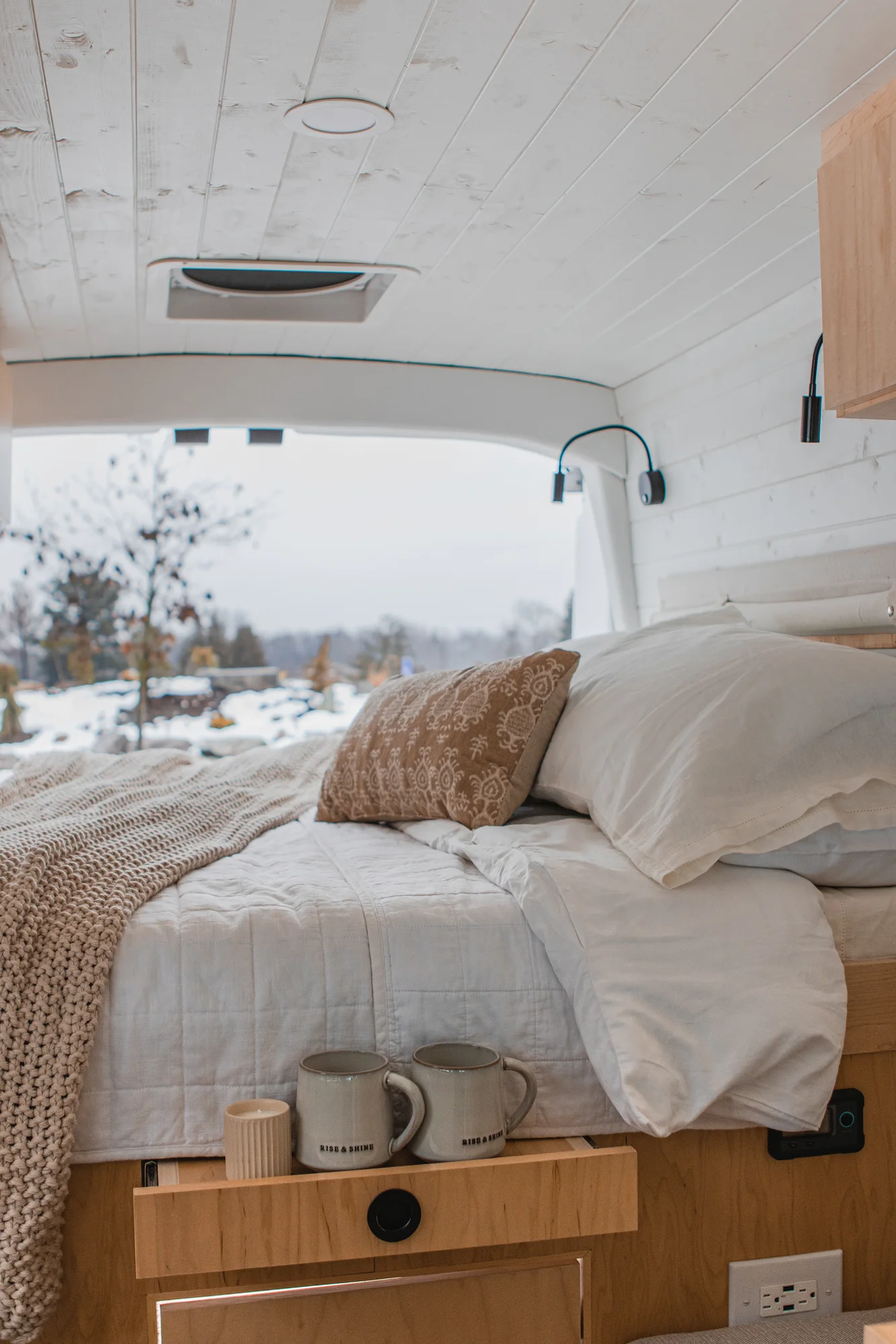 The Drifter 101 custom Ford Transit campervan built for Zach and Jacqueline by Drifter Vans, with a matte black sink and faucet, light wood cabinetry, and a raised bed with overhead shelves