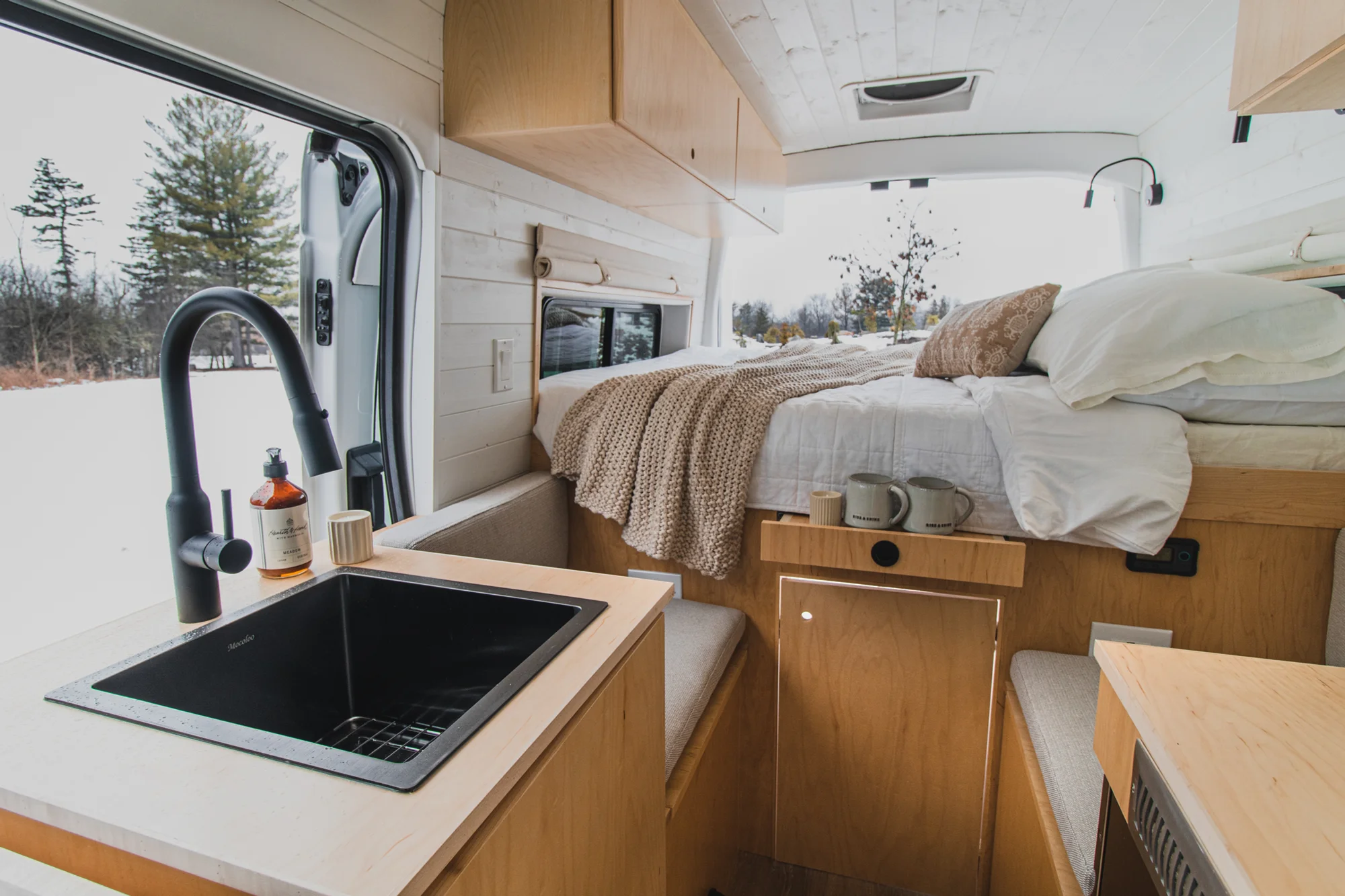 The Drifter 101 custom Ford Transit campervan built for Zach and Jacqueline by Drifter Vans, with a matte black sink and faucet, light wood cabinetry, and a raised bed with overhead shelves