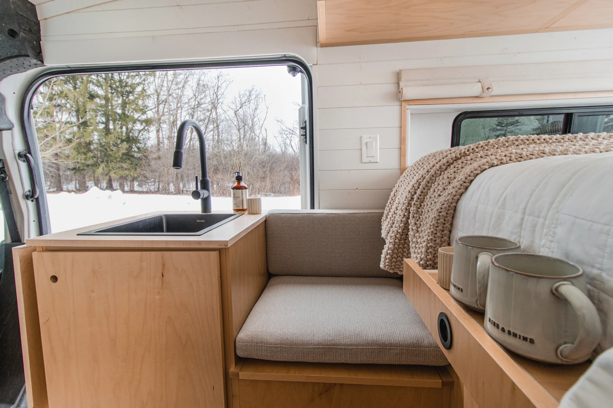 The Drifter 101 custom Ford Transit campervan built for Zach and Jacqueline by Drifter Vans, with a matte black sink and faucet, light wood cabinetry, and a raised bed with overhead shelves