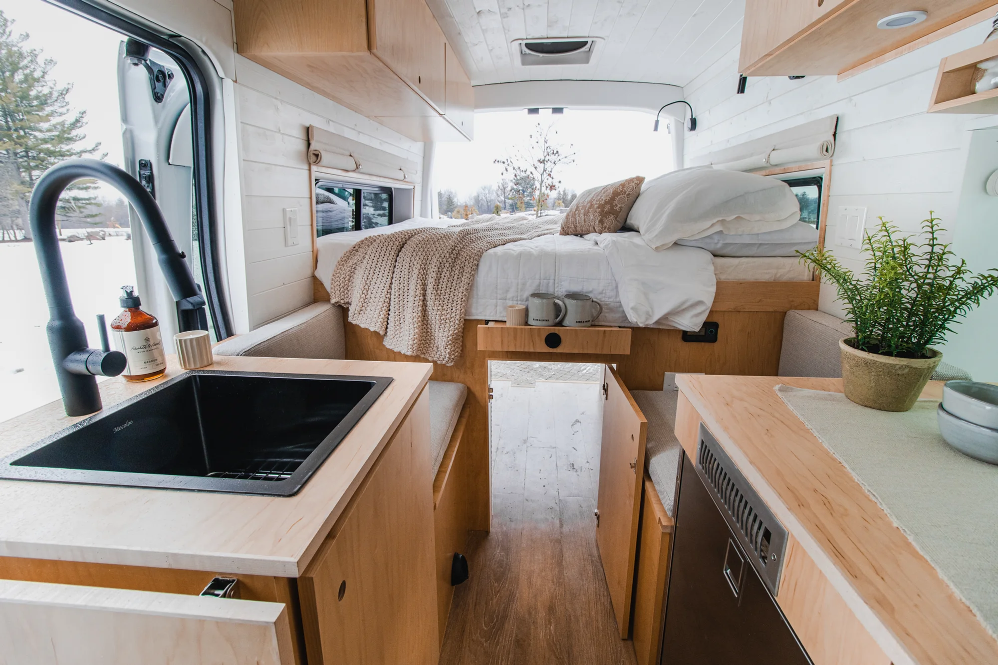 The Drifter 101 custom Ford Transit campervan built for Zach and Jacqueline by Drifter Vans, with a matte black sink and faucet, light wood cabinetry, and a raised bed with overhead shelves