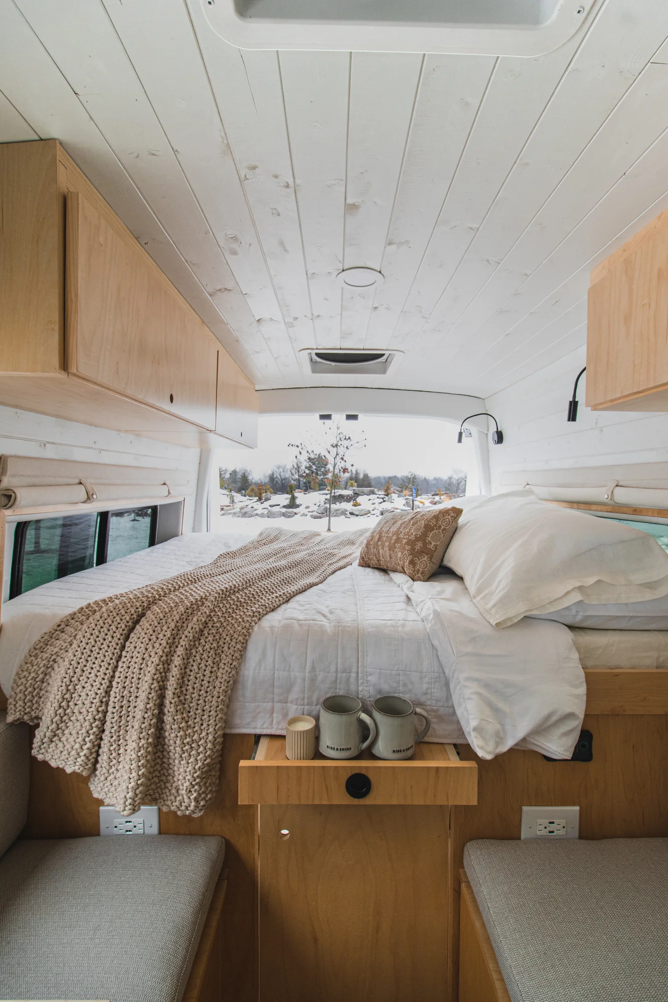 The Drifter 101 custom Ford Transit campervan built for Zach and Jacqueline by Drifter Vans, with a matte black sink and faucet, light wood cabinetry, and a raised bed with overhead shelves