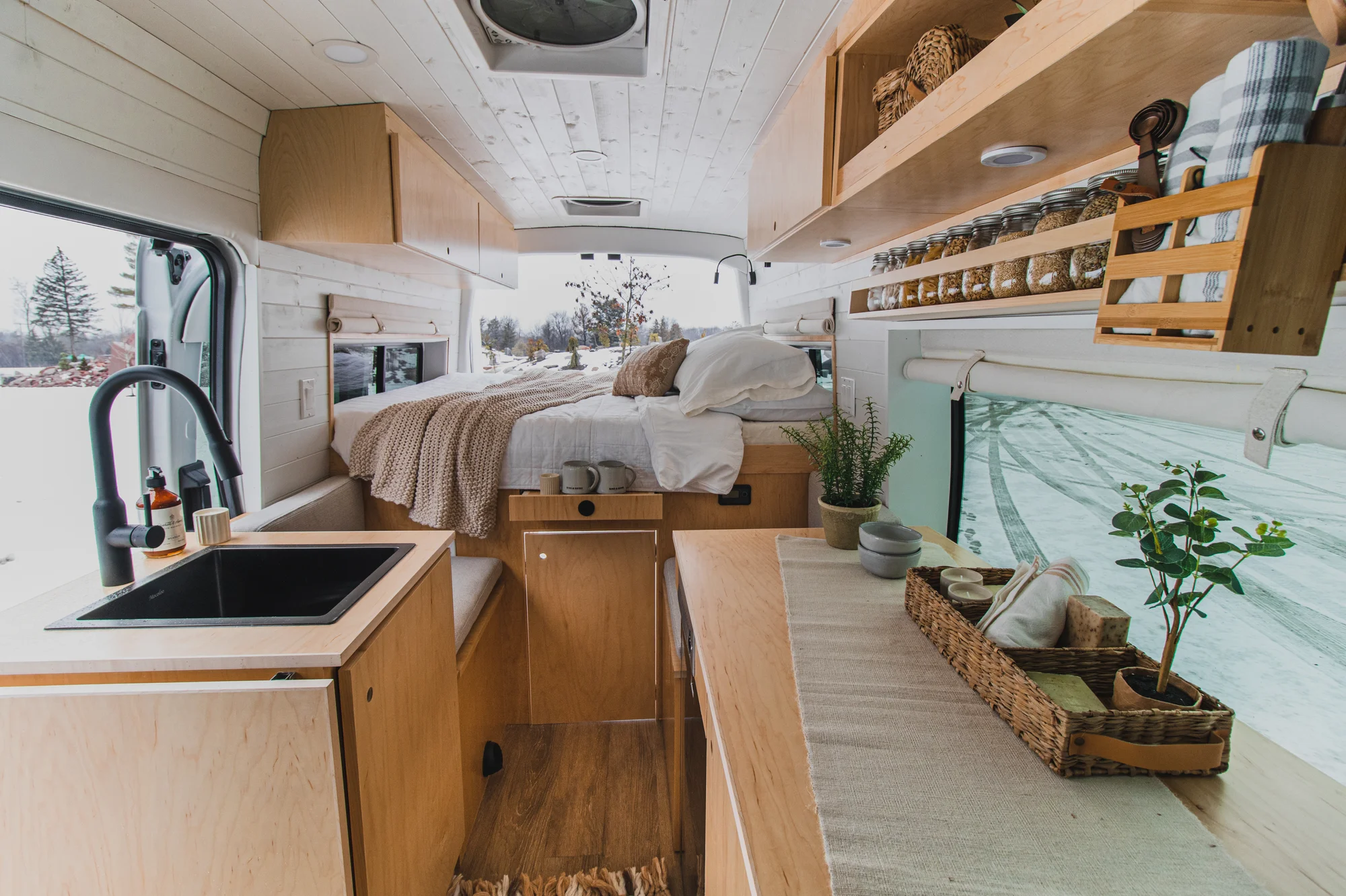The Drifter 101 custom Ford Transit campervan built for Zach and Jacqueline by Drifter Vans, with a matte black sink and faucet, light wood cabinetry, and a raised bed with overhead shelves