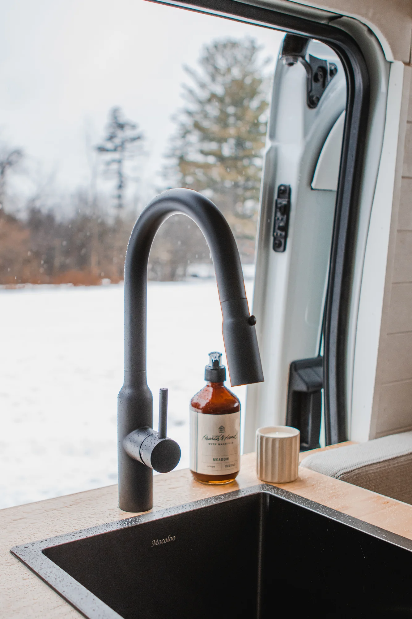 The Drifter 101 custom Ford Transit campervan built for Zach and Jacqueline by Drifter Vans, with a matte black sink and faucet, light wood cabinetry, and a raised bed with overhead shelves