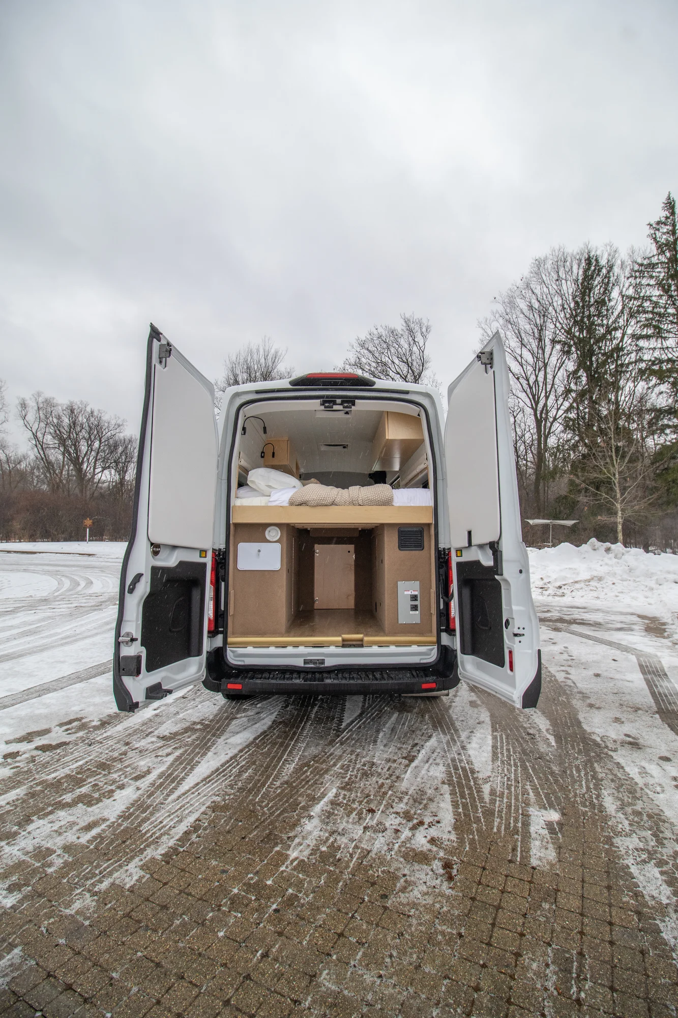The Drifter 101 custom Ford Transit campervan built for Zach and Jacqueline by Drifter Vans, with a matte black sink and faucet, light wood cabinetry, and a raised bed with overhead shelves