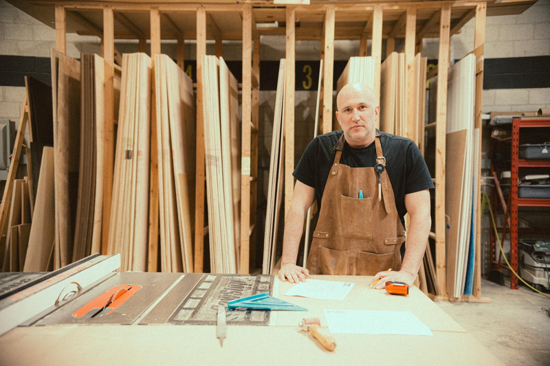Paul Domish leaning casually on a wooden workbench at Drifter Vans HQ, surrounded by tools, campervan components, and soft overhead lighting—capturing a moment of calm in the creative process.