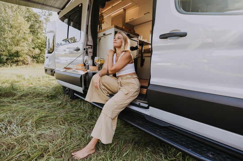 Beautiful blond woman sitting on the edge of her transit camper van looking out with a smile