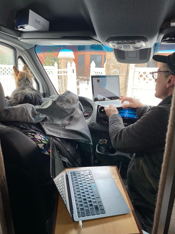 Roger working from the front seat of the van, parked at a ski resort with snow-covered mountains visible in the background—blending remote work with adventure.