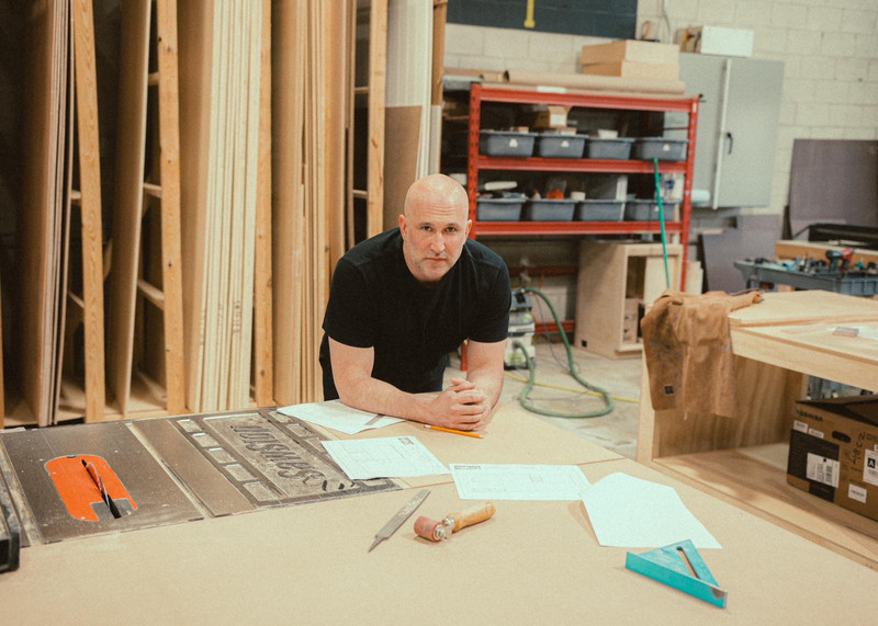 Paul Domish at Drifter Vans HQ wearing a work apron, standing in the workshop surrounded by tools and custom van components.