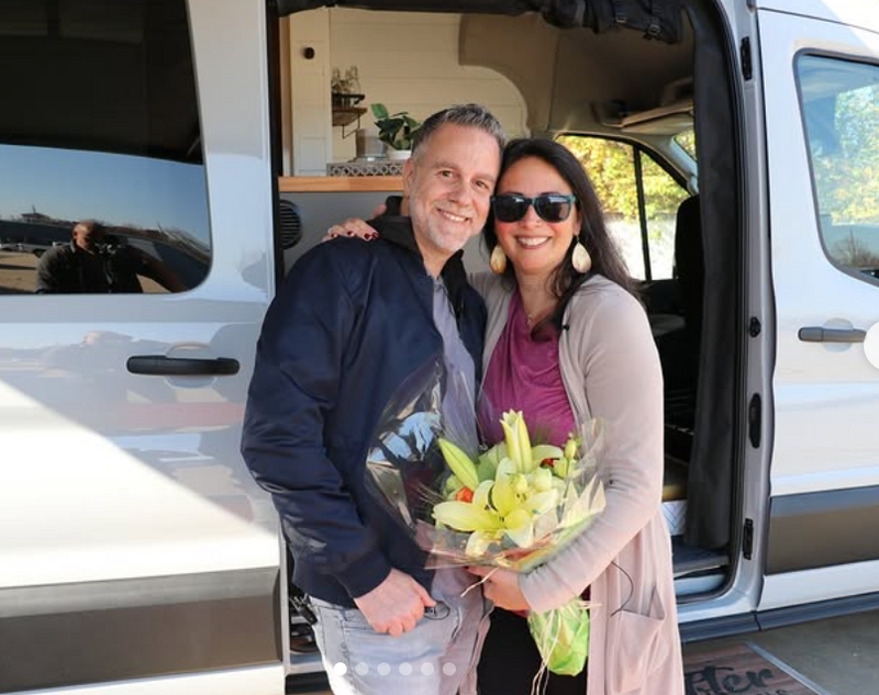 Couple standing in front of their newly built camper van holding fresh flowers, celebrating the start of their vanlife journey.