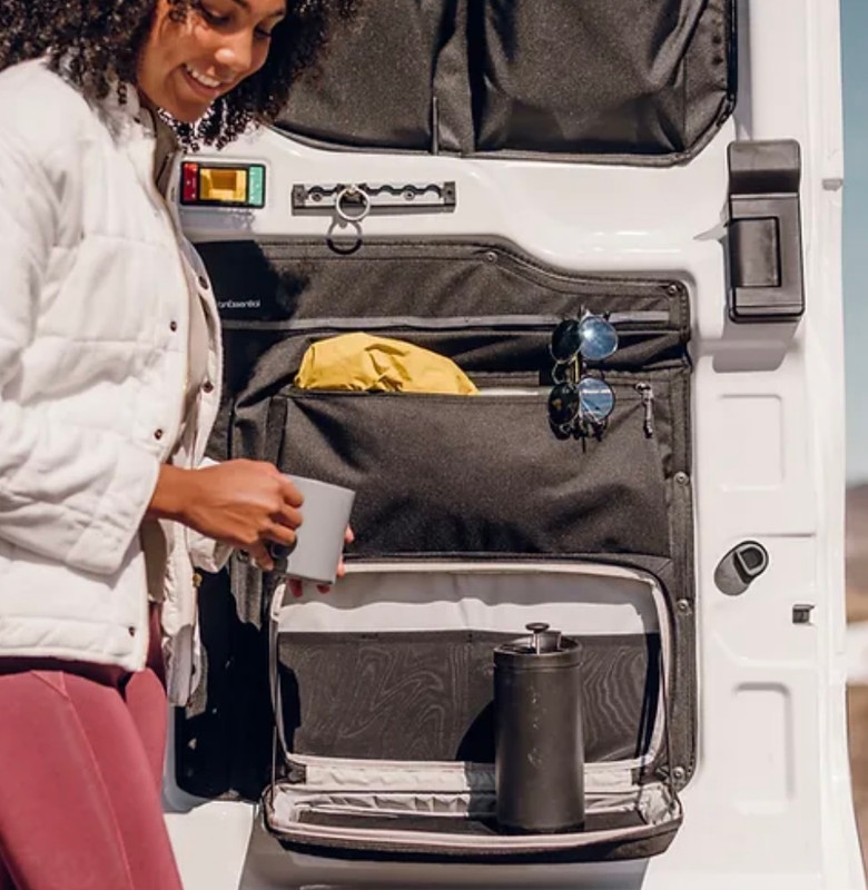 Woman standing beside rear doors of a Ford Transit campervan, showcasing VanEssential rear door storage bags for vanlife organization