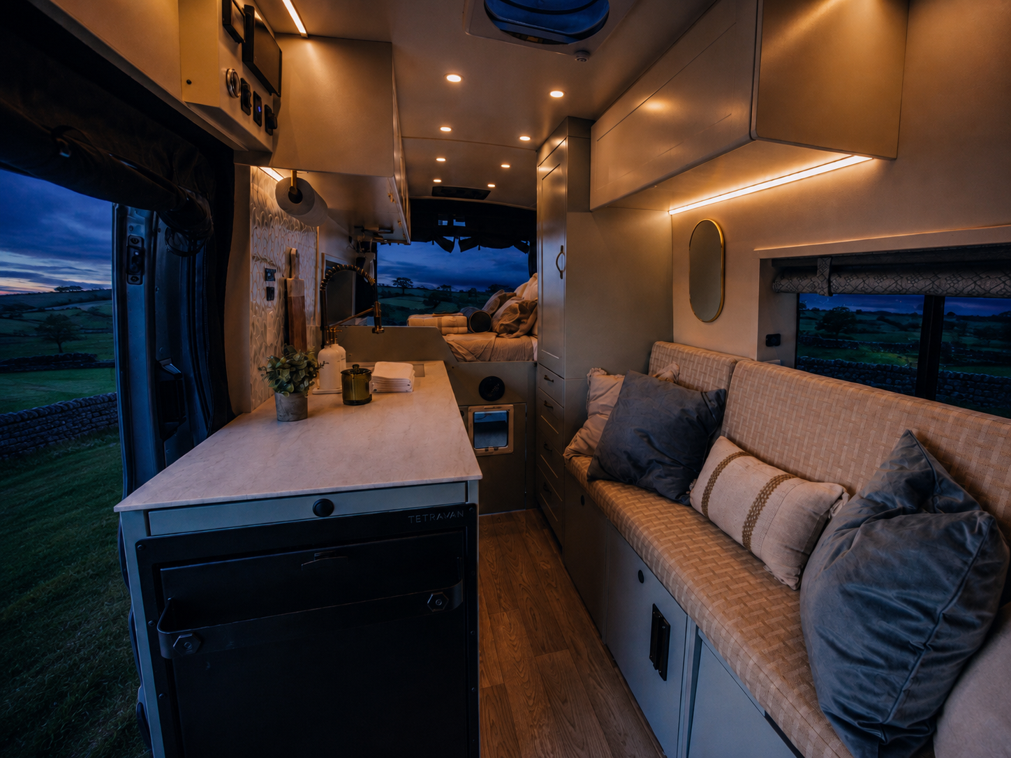 The Drifter 101 custom Ford Transit campervan built for Kathy by Drifter Vans, Drifter layout with Wedgewood Grey cabinetry, a Beige Travertine countertop, vintage brass faucet, and Charlotte Rattan cushions