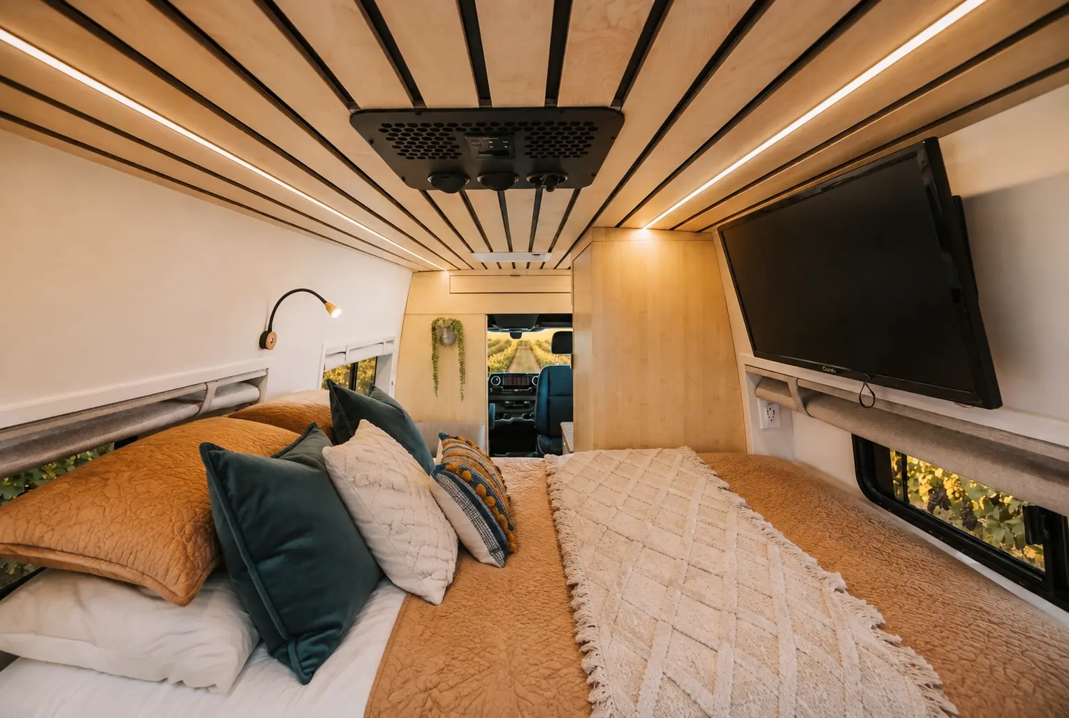 The Drifter 101 Mercedes-Benz Sprinter campervan built for Rosa by Drifter Vans: full bed platform with earth-tone bedding, wall-mounted TV, and natural bamboo slat ceiling looking toward the cab