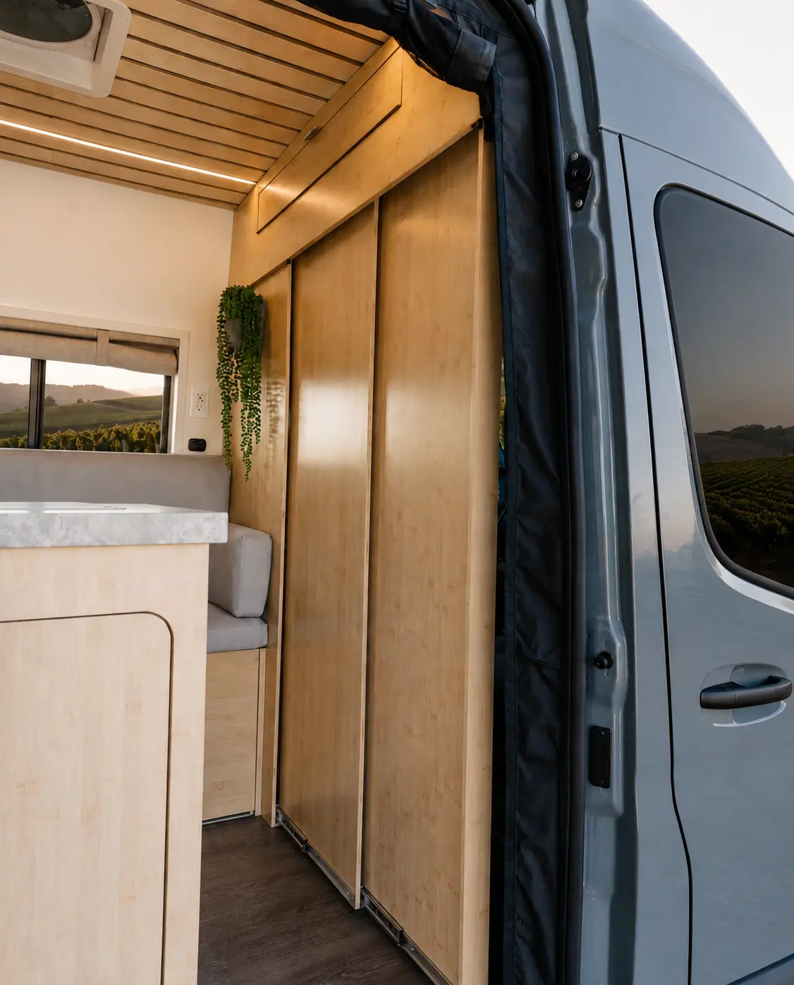 The Drifter 101 Mercedes-Benz Sprinter campervan built for Rosa by Drifter Vans: view through the open sliding door showing bamboo wardrobe cabinets, stone-look counter and bench seating, vineyard at sunset