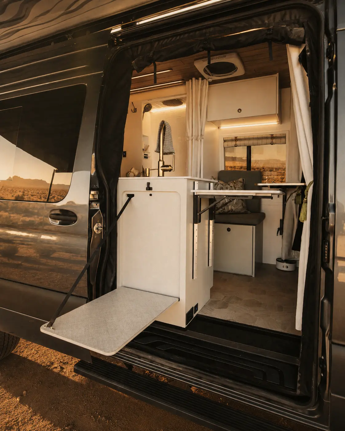 The Drifter 101 Mercedes-Benz Sprinter Coachcraft campervan built for Trish by Drifter Vans: sliding door open with fold-out exterior table, brass faucet galley, dinette and shower beyond, desert sunset reflected on the van