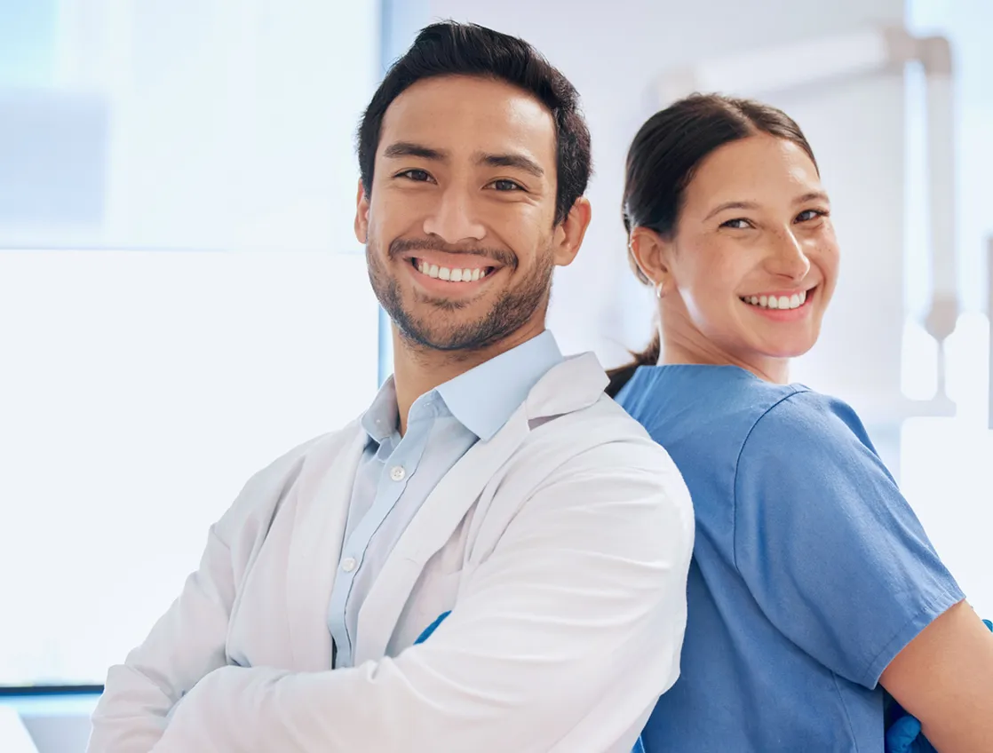 A man and a woman, both smiling, stand back to back, wearing medical attire.