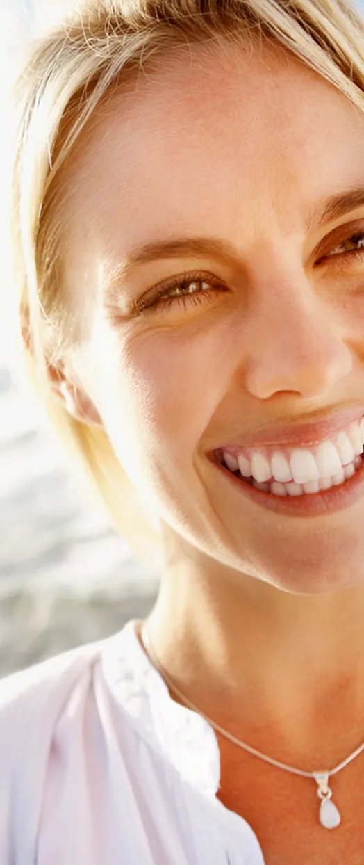 A woman with blonde hair smiles brightly, wearing a white top and necklace.