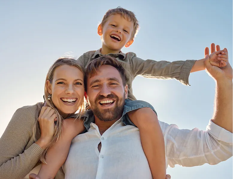 A smiling family, with a child sitting on the father's shoulders, enjoying a sunny day.