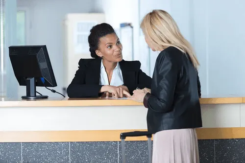 A receptionist talks to a woman standing at a desk with a suitcase handle visible.