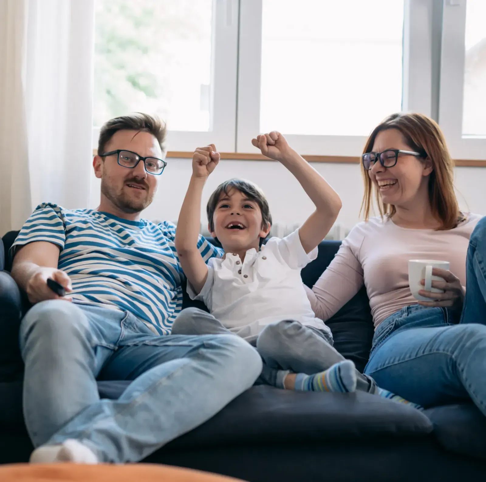 A family of three sits on a couch, smiling and cheering while watching TV.