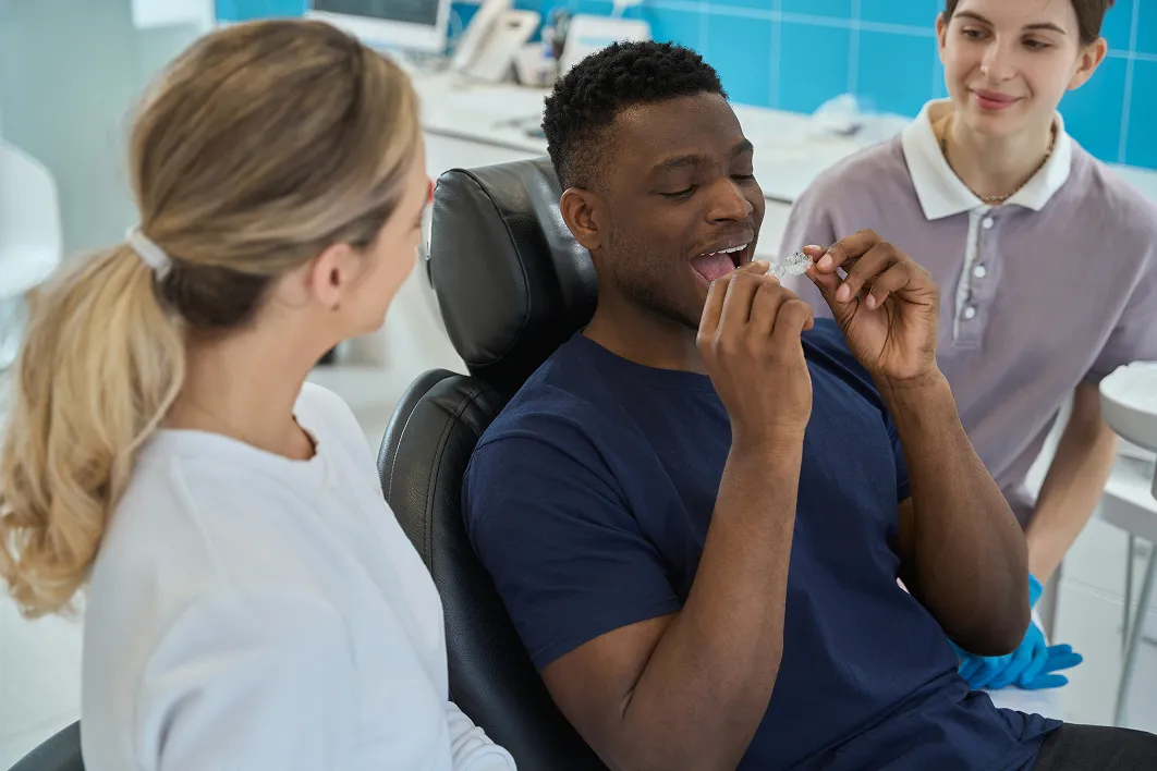 Man in dental chair examines a clear aligner, two dental professionals assist nearby.