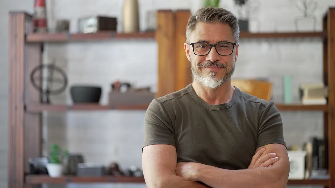 A man with glasses and a grey beard smiles with arms crossed in front of shelves.