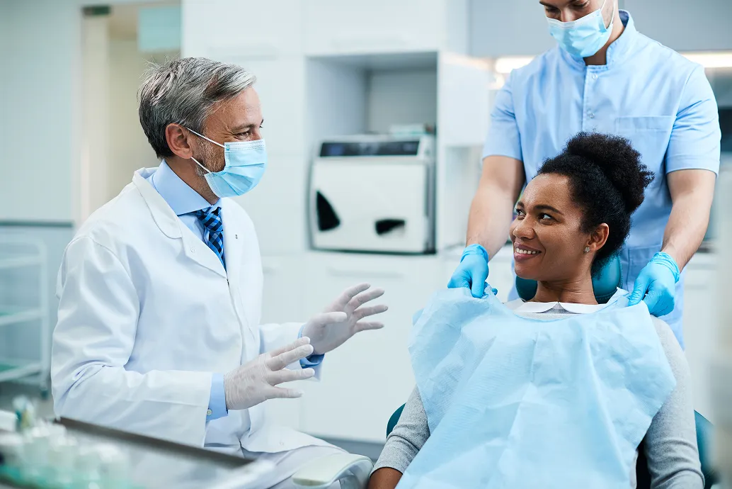 A dentist talks to a smiling patient in a dental chair, assisted by another person.