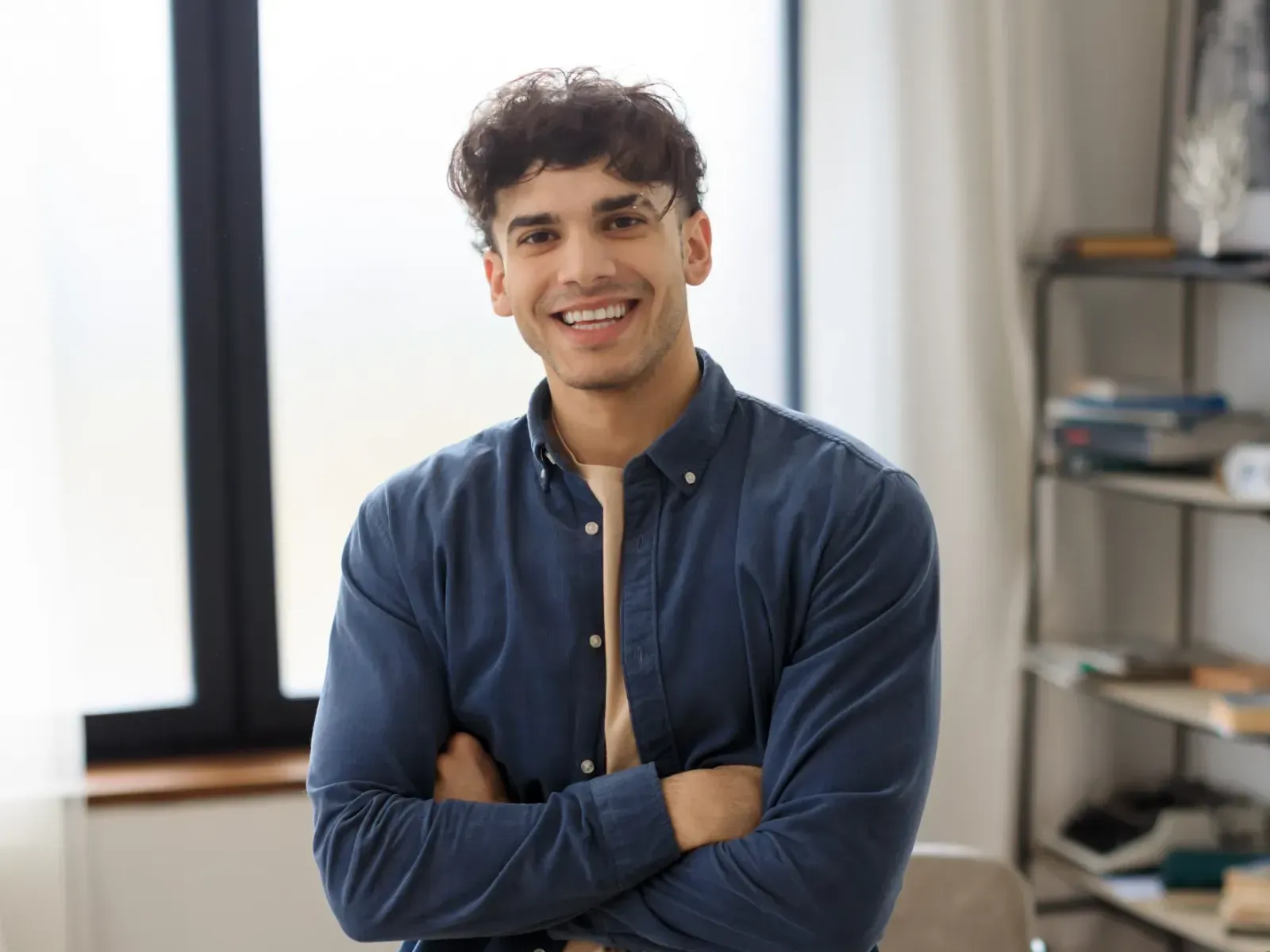 A person in a blue shirt smiles with arms crossed, standing indoors near a window.