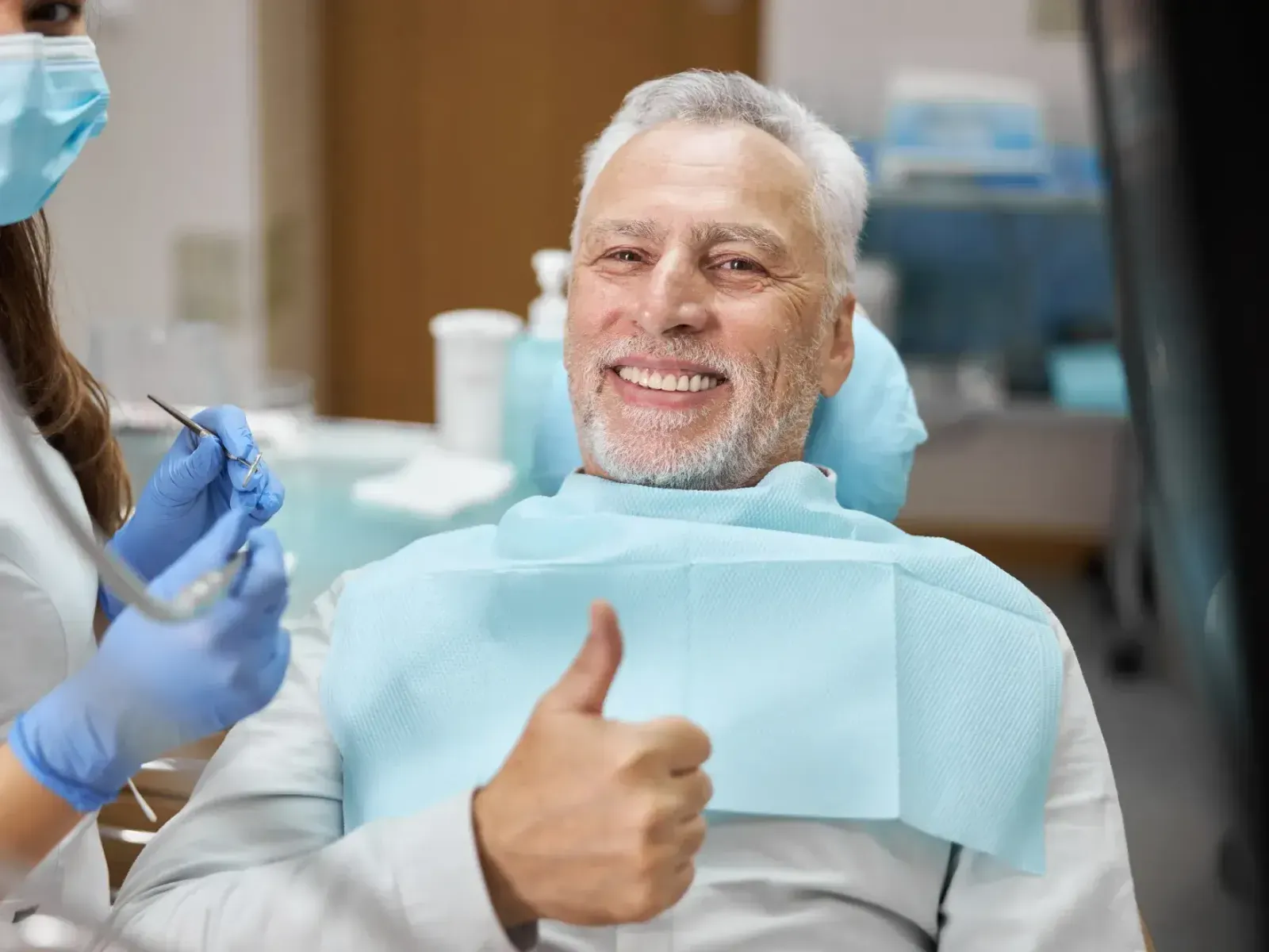Smiling man in dental chair giving thumbs up, dental assistant holding dental tools beside him.