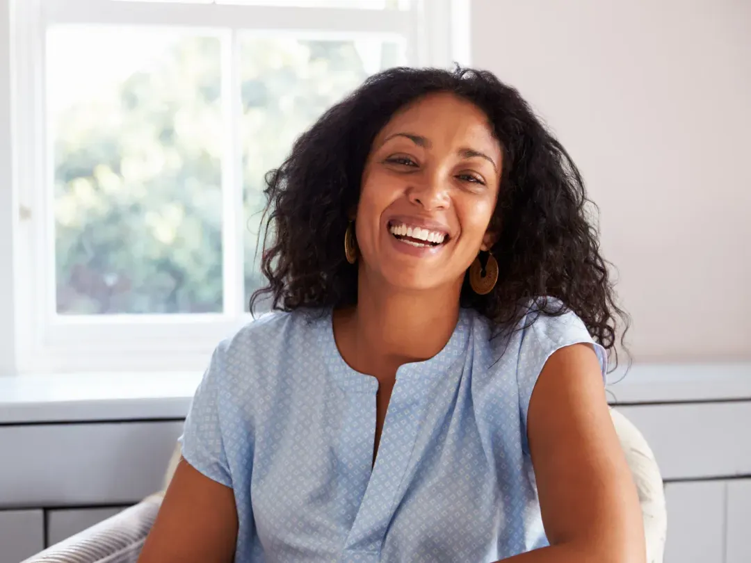 A woman with curly hair is smiling indoors, sitting in front of a window.