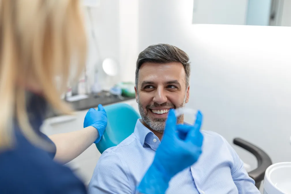 A dentist shows a dental tool to a smiling man sitting in a dental chair.
