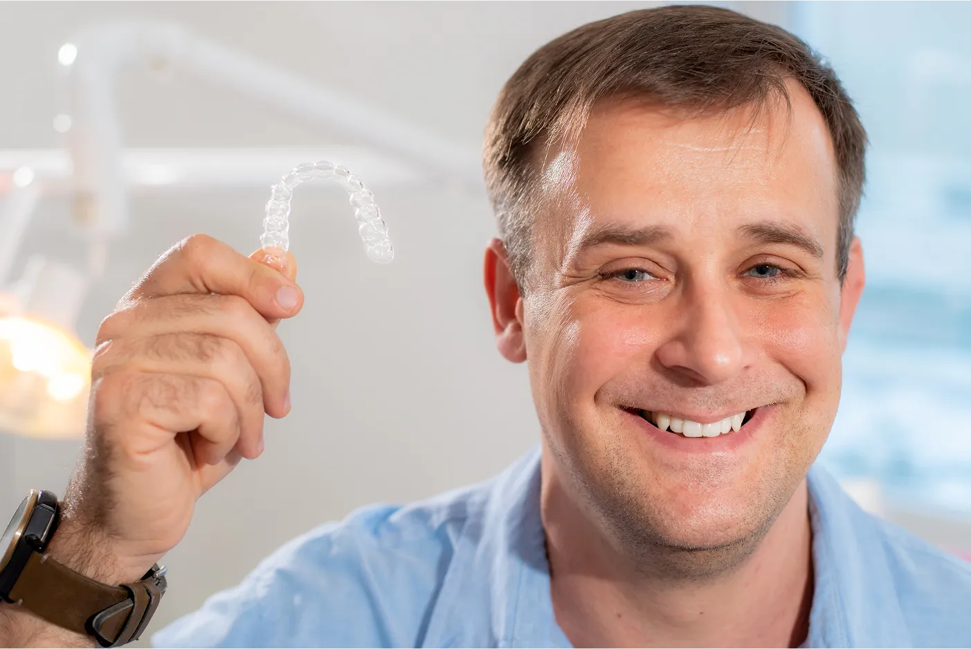 A man smiling, holding a clear dental aligner with his right hand.