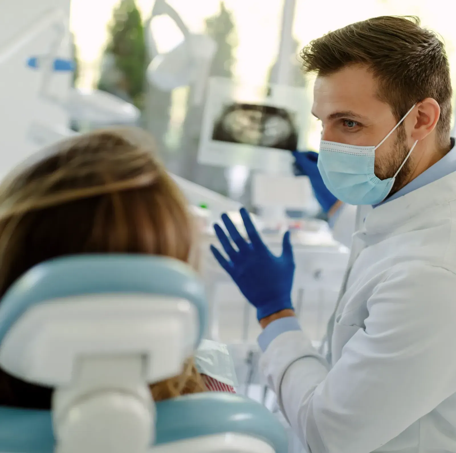 Dentist wearing a mask and gloves discusses X-ray results with a patient in a dental office.