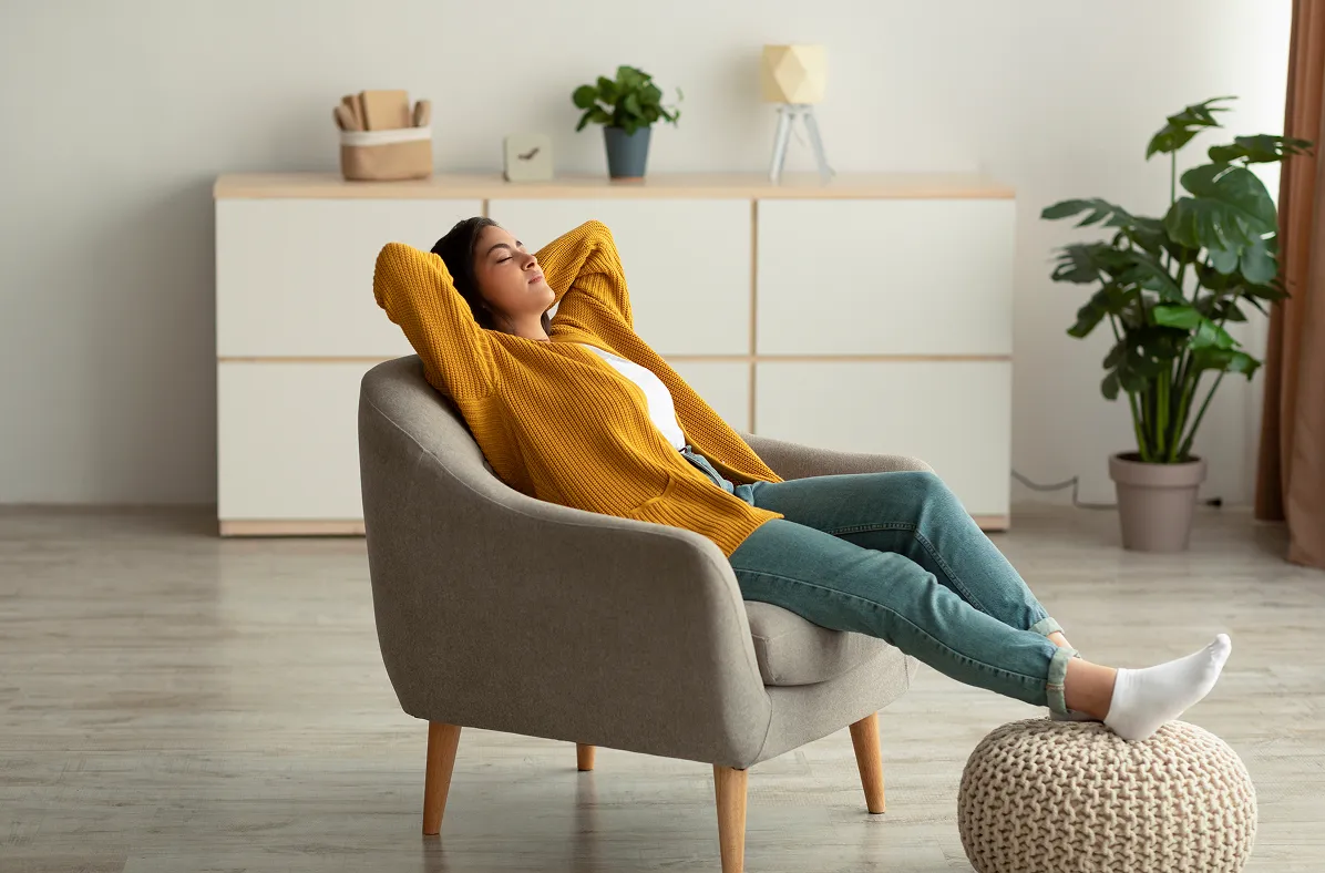 Person relaxing in a chair with feet up on a footstool, eyes closed, looking content.