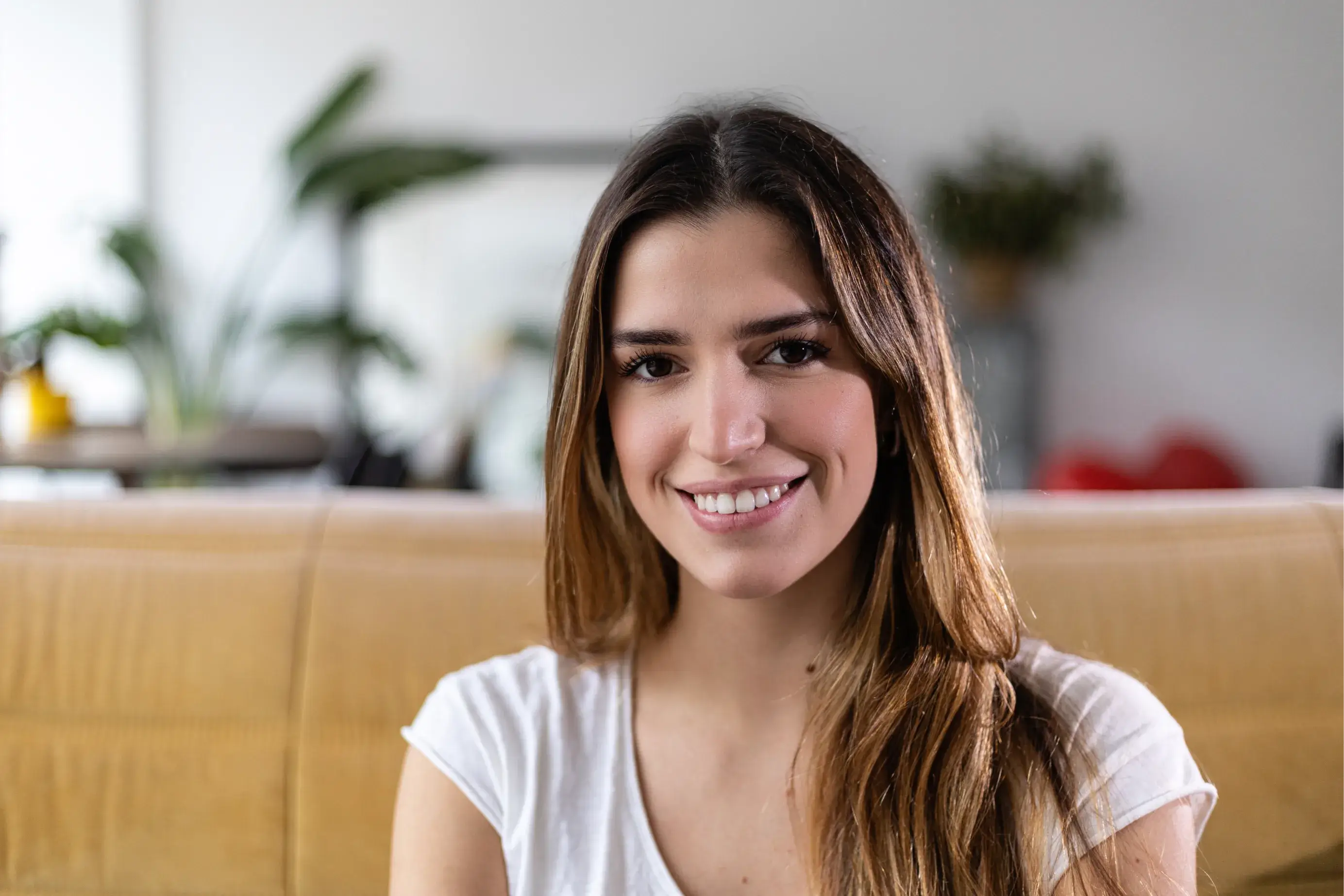 Smiling woman sitting on a couch, wearing a white shirt, with plants in the background.