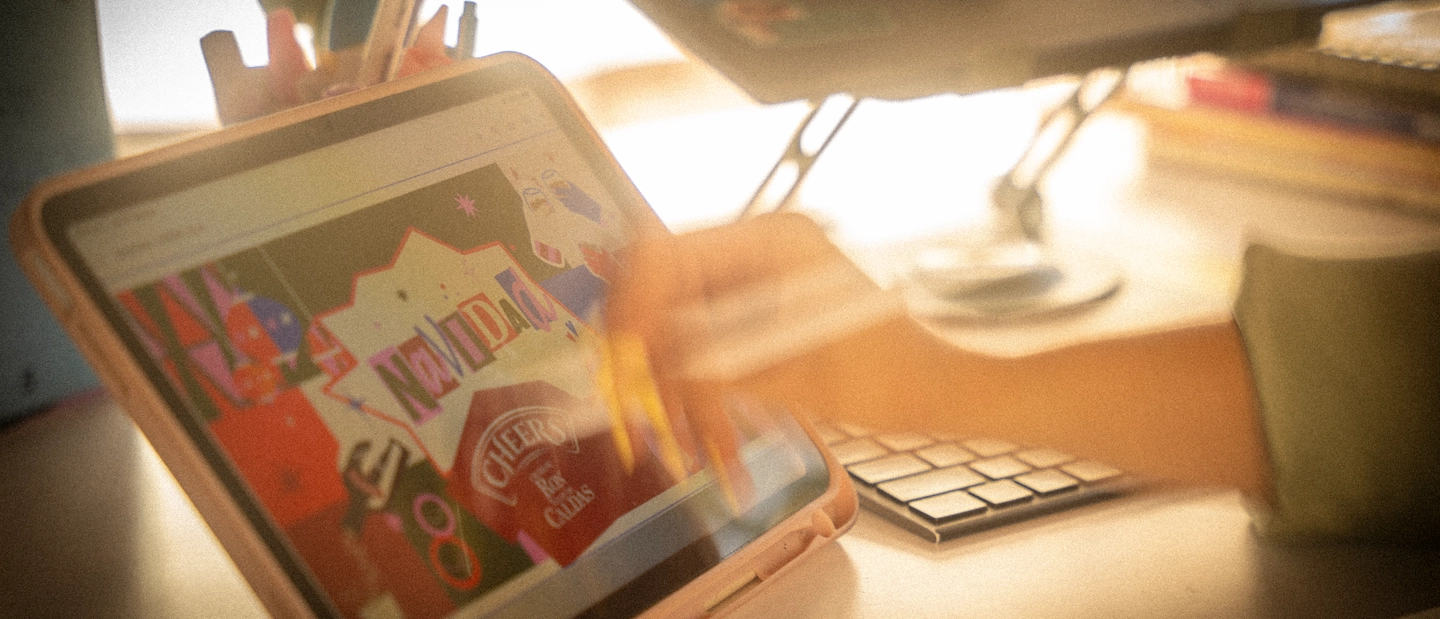 Tablet sobre un escritorio de trabajo creativo mostrando en pantalla un diseño gráfico navideño con la palabra 'Navidad', iluminada por luz cálida. Al lado se ven un teclado, un portalápices y utensilios de diseño, con desenfoque que evoca un ambiente de estudio en pleno proceso creativo.