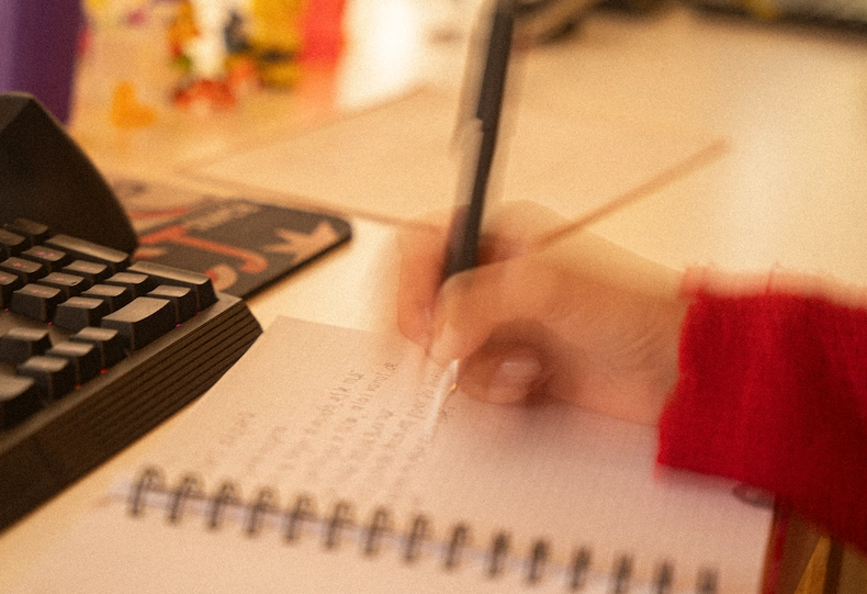 Mano escribiendo a mano con bolígrafo en un cuaderno de espiral sobre un escritorio de trabajo con teclado, en luz cálida y con leve desenfoque que evoca un momento genuino de ideación creativa.
