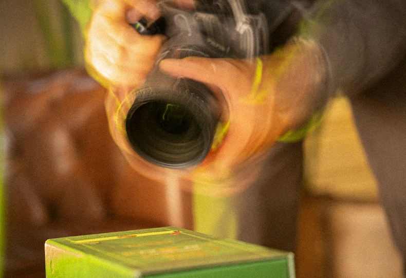 Persona fotografiando de cerca una caja verde de producto con una cámara réflex, con desenfoque de movimiento y luz cálida que refleja un cuidadoso trabajo de fotografía de producto.