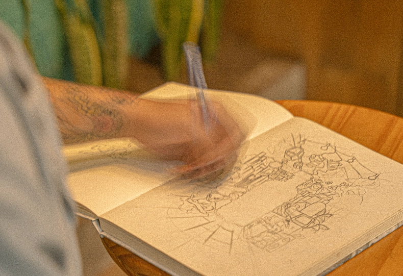 Persona con tatuaje en el brazo dibujando bocetos a lápiz en un cuaderno apoyado sobre una mesa de madera, junto a una planta, en ambiente cálido que transmite un proceso creativo de ilustración.