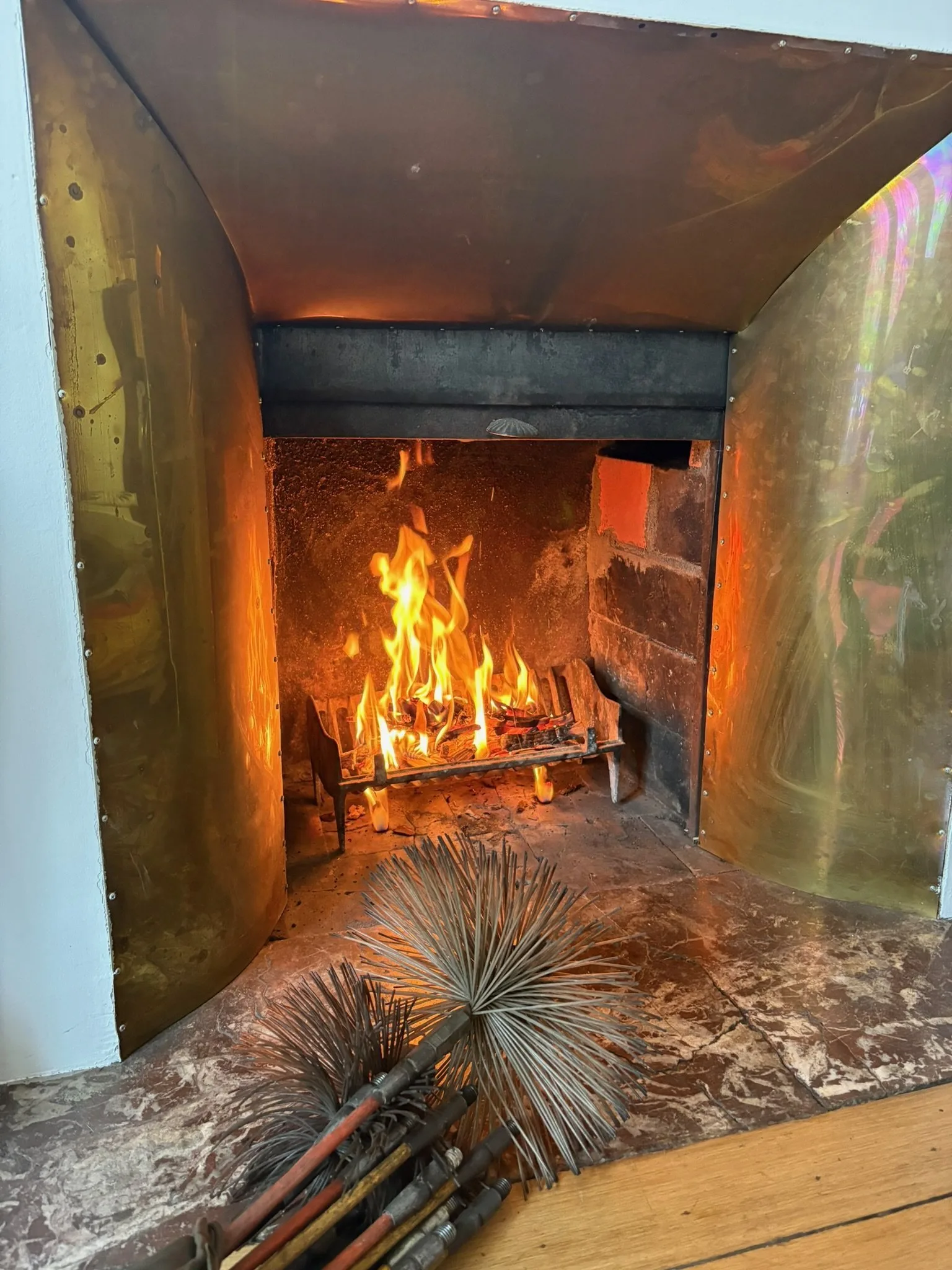 Feu allumé dans une cheminée à hotte laiton, cannes et hérisson de ramonage posés sur l'âtre