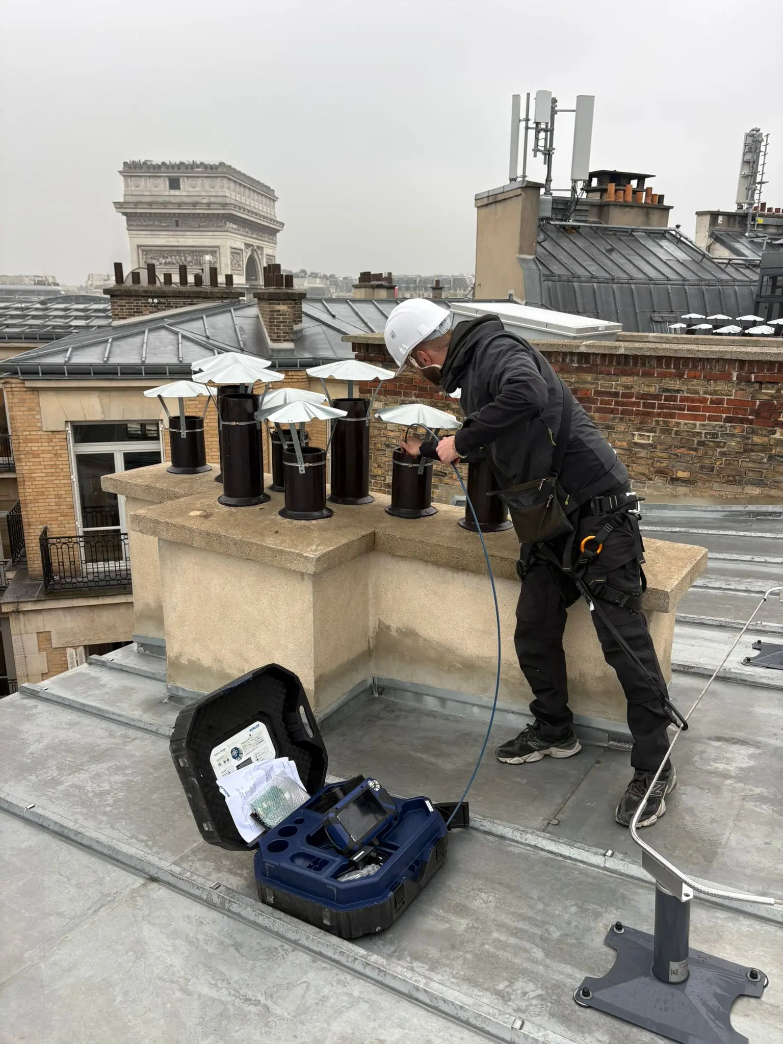 Diagnostic de conduits sur une toiture parisienne, technicien harnaché et mallette d'inspection au sol