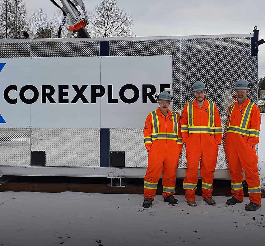 Four Corexplore crew members in orange coveralls and hard hats outside a branded Corexplore drill shack in winter
