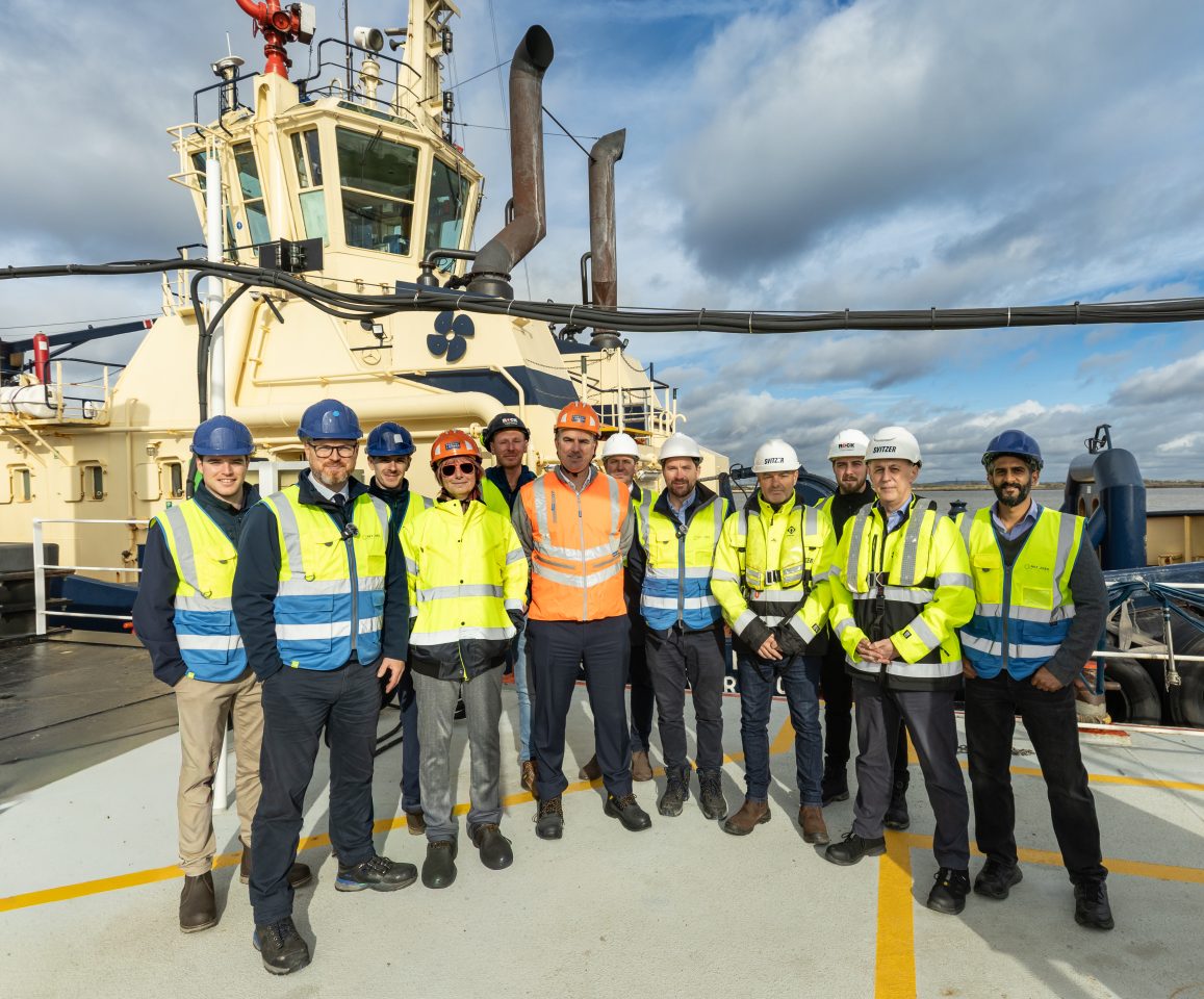Group of twelve workers in safety gear standing on a ship deck with a ship cabin in the background.