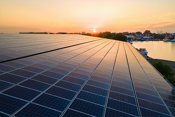 Large solar panel array at sunset near a river with boats and buildings in the background.