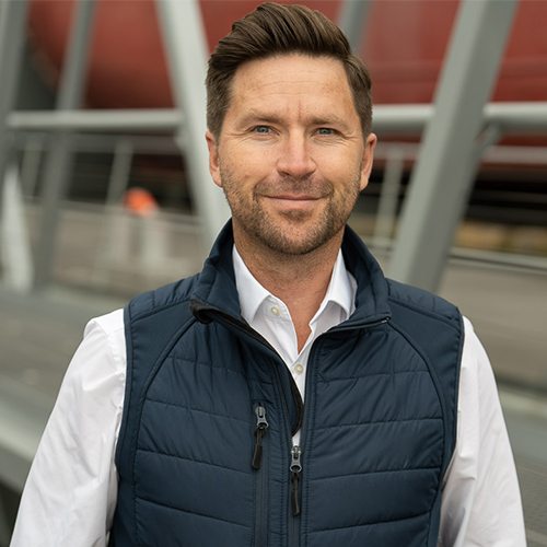 Smiling man with neatly styled hair wearing white shirt and navy blue quilted vest outdoors.
