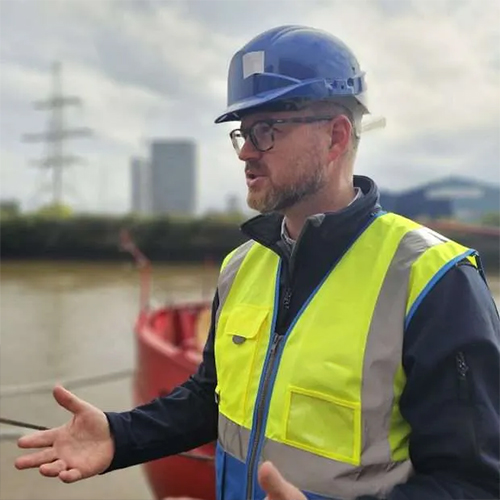 Man wearing blue hard hat and yellow safety vest gesturing near water with buildings in background.
