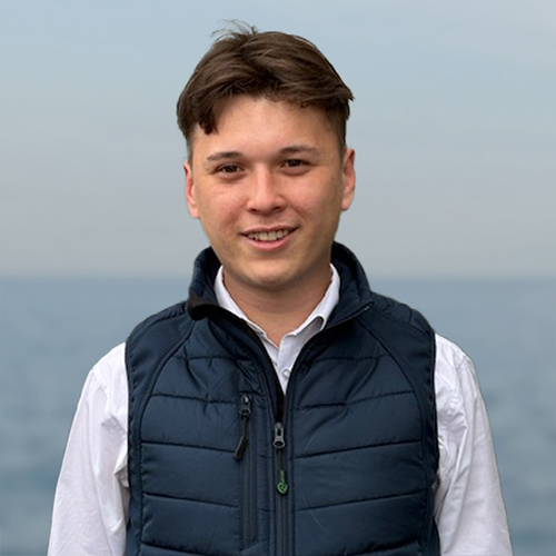 Smiling young man wearing a blue vest over a white shirt with a blurred ocean background.