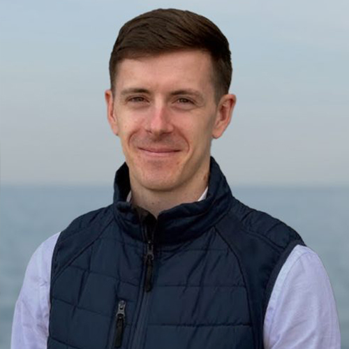 Smiling man with short brown hair wearing a navy vest over a white shirt with a sea background.