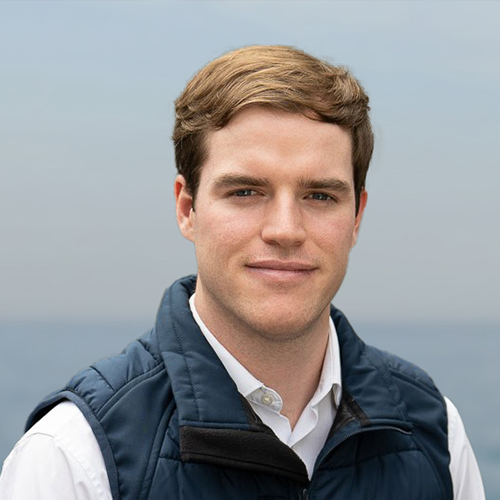 Portrait of a young man with short brown hair wearing a white shirt and navy blue vest outdoors.