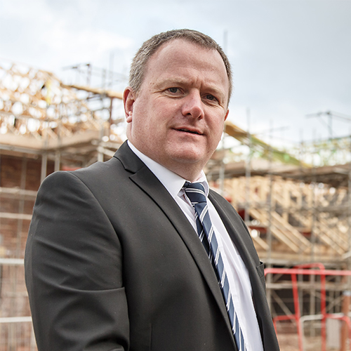 Man in a suit and striped tie standing in front of a construction site with scaffolding.