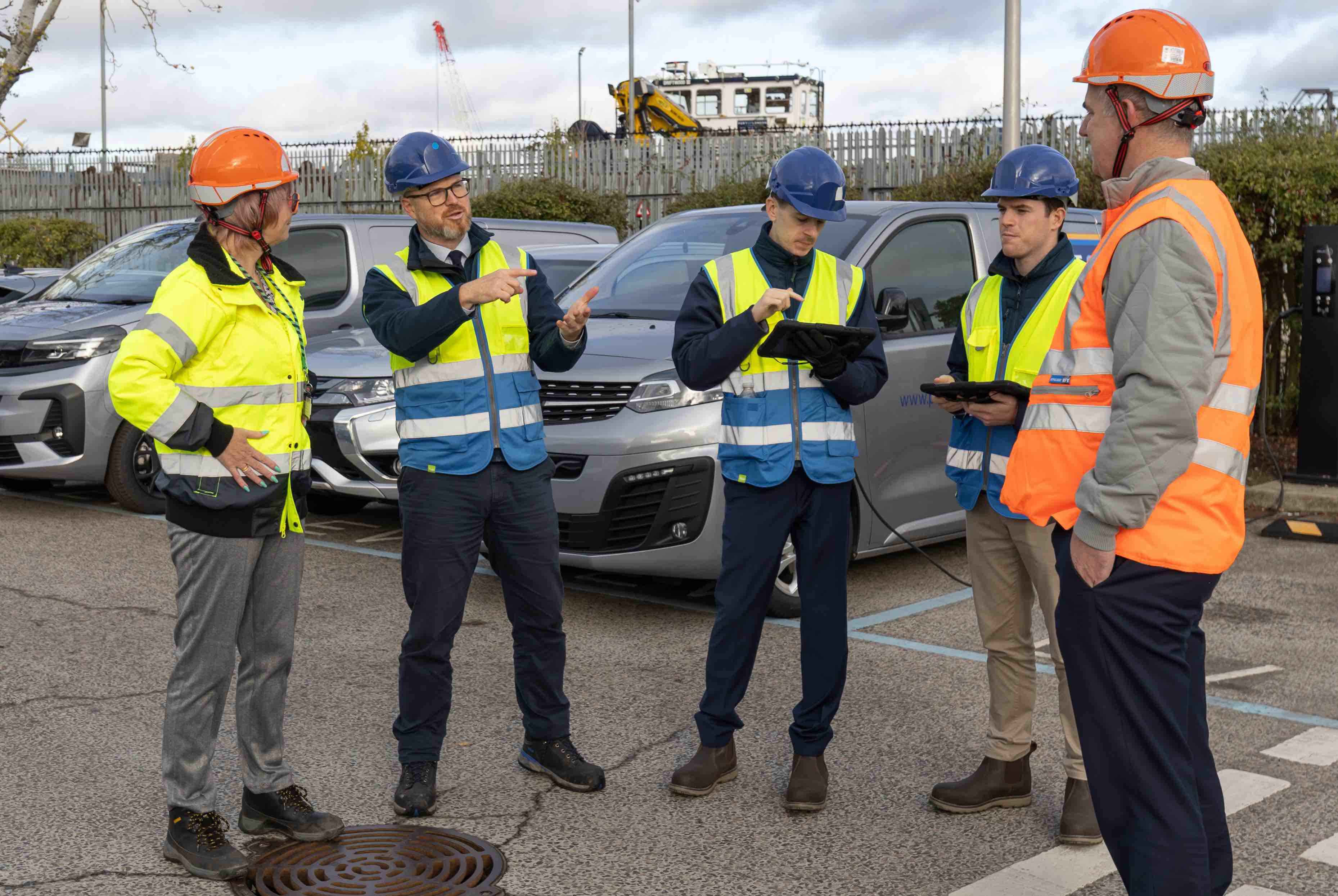 Construction workers in safety vests and helmets discussing plans outdoors near parked vehicles.